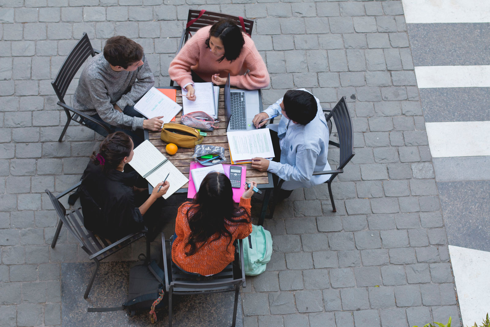 Grupo de cinco estudiantes estudiando con apuntes y computador, sentados en una mesa del patio